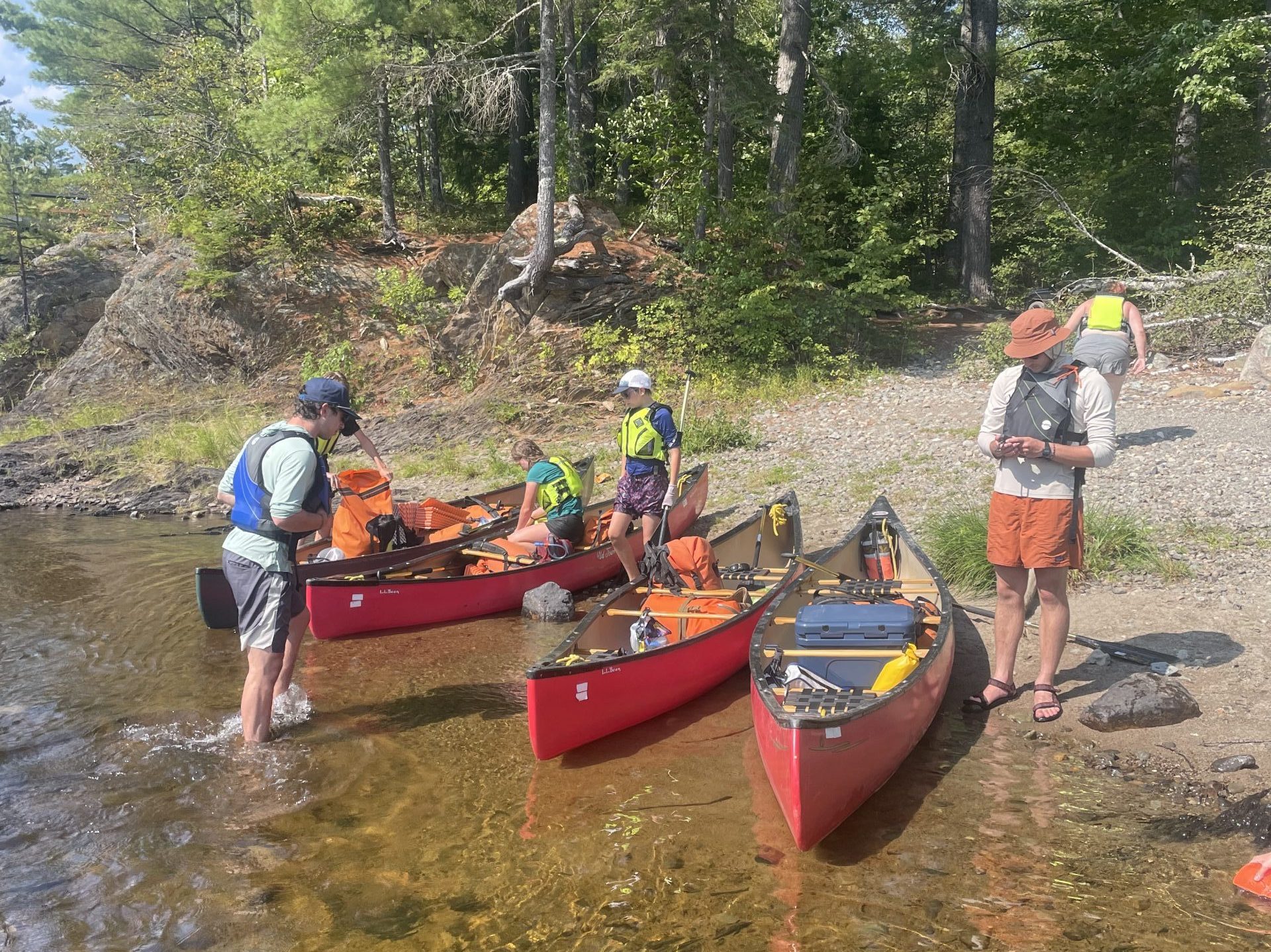 People preparing canoes on a shore, surrounded by trees, on a sunny day.