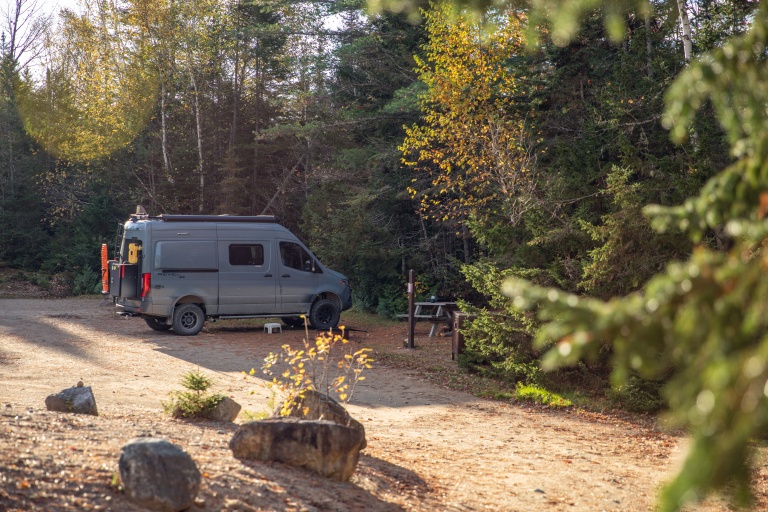 A gray van parked in a forest clearing with colorful autumn leaves and picnic table nearby.