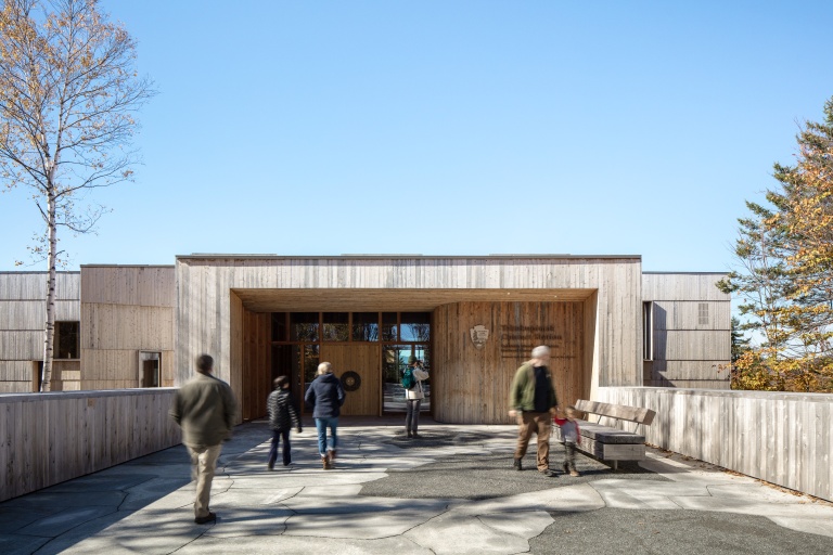 Modern wooden building entrance with visitors walking on a stone pathway, surrounded by trees and blue sky.