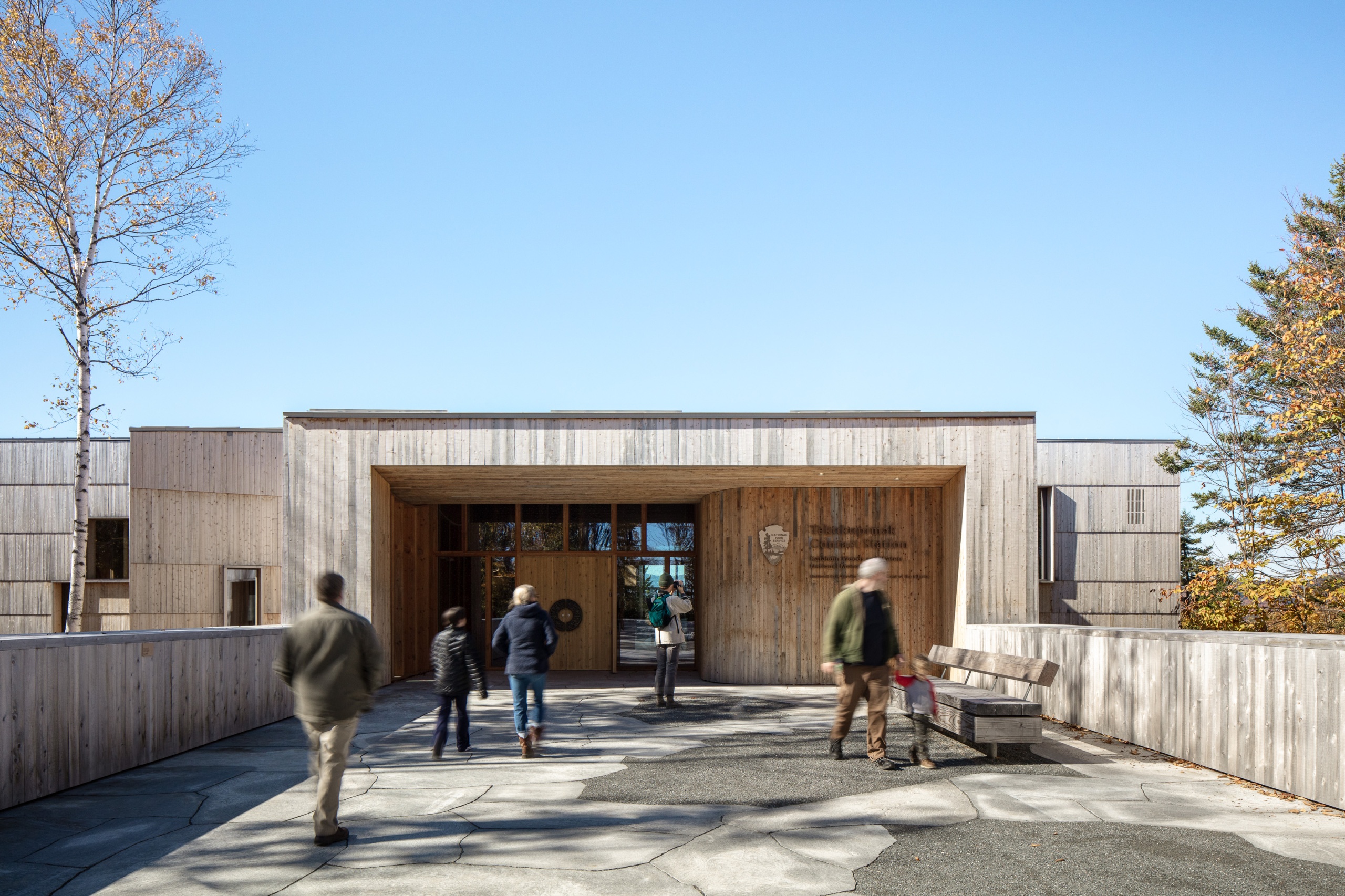 Modern wooden building entrance with visitors walking on a stone pathway, surrounded by trees and blue sky.