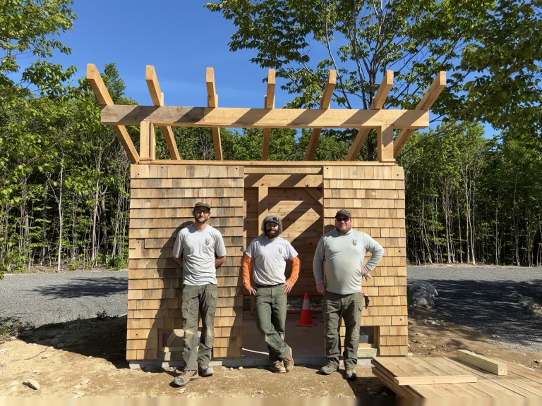 Three individuals stand in front of a partially constructed wooden structure under a clear blue sky.