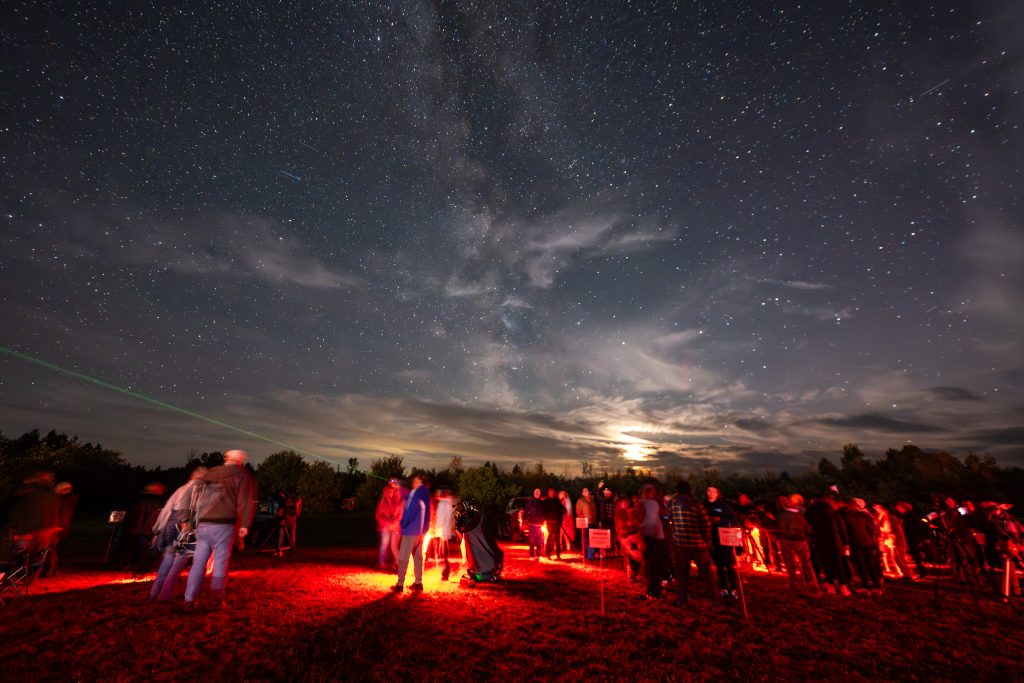 A starry night sky filled with the Milky Way, with a crowd observing and a green laser pointing upwards.