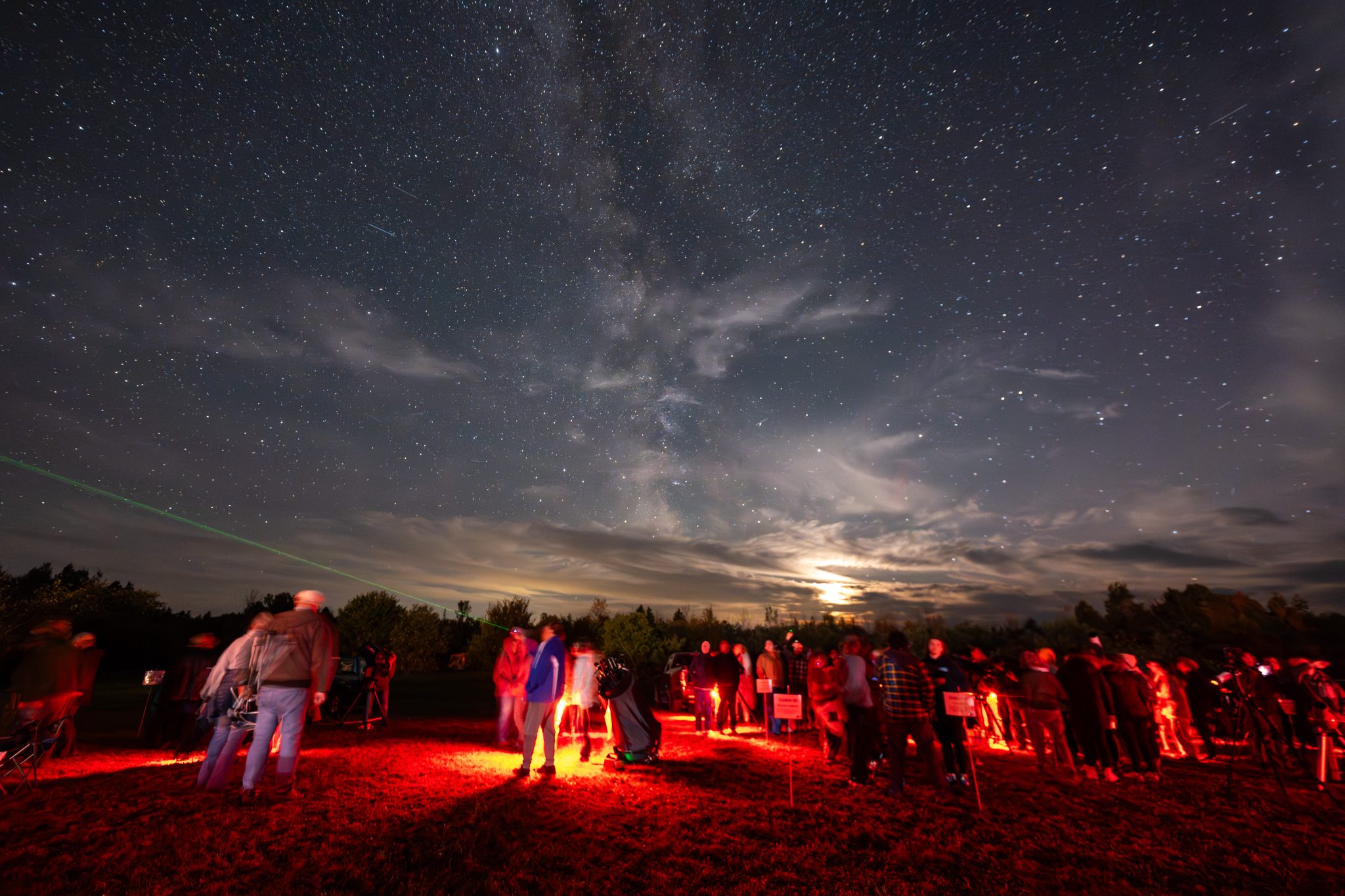A starry night sky filled with the Milky Way, with a crowd observing and a green laser pointing upwards.