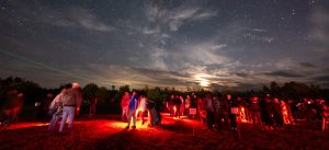 A starry night sky filled with the Milky Way, with a crowd observing and a green laser pointing upwards.