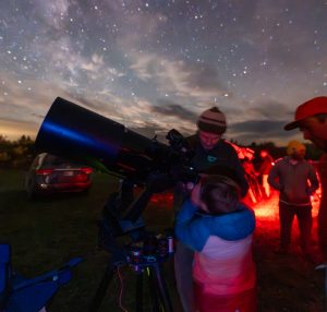 A child looks through a telescope, surrounded by adults under a starry sky, with red-lit surroundings.