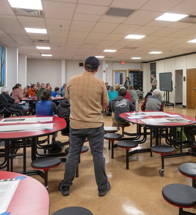 A man stands in front of a meeting where a group of people are seated, discussing topics in a community space.