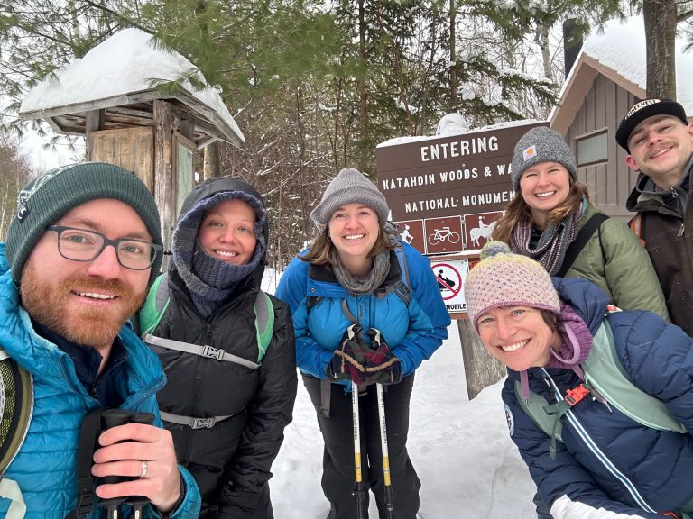 A group of six friends poses for a selfie in the snowy woods near the Katahdin Woods & Waters sign.