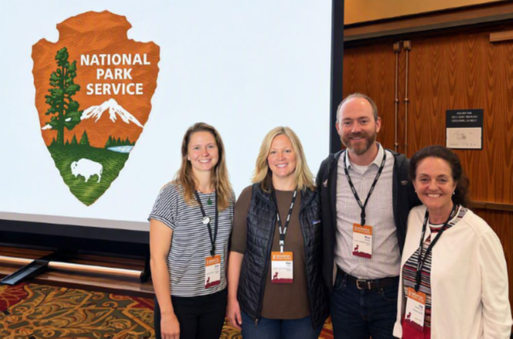 4 staff members standing together at the National Park Friends Alliance annual meeting in front of an NPS presentation slideshow.