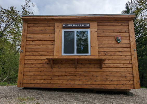 A small clapboard shed with natural stain bears the NPS arrowhead logo and a nameplate: Katahdin Woods and Waters.