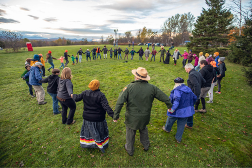 A circle comprising about 35 people are joined hand to hand in a grassy field.