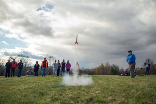 A model rocket ship is 6 feet from the ground with a puff of smoke below. A group of teens are gathered around.