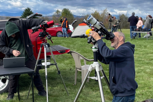 Two white men look through telescopes during the day in a grassy field.