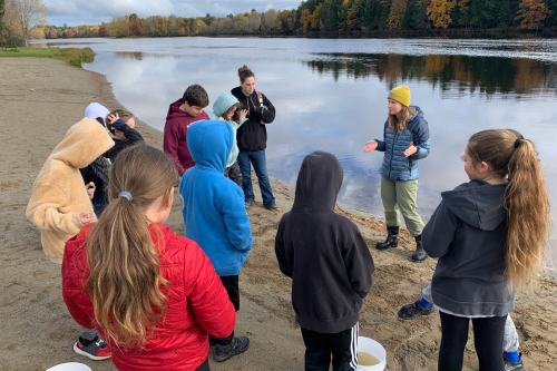 Six elementary students and two adult women stand on a sandy river beach.