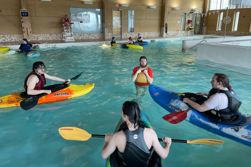 High school students sitting in kayaks and an instructor standing in an indoor swimming pool.