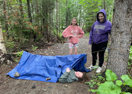 Two middle school girls stand on a trail, one middle school boy is in an improvised tarp tent.