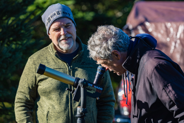 A man watches another man looking through a telescope in daylight.