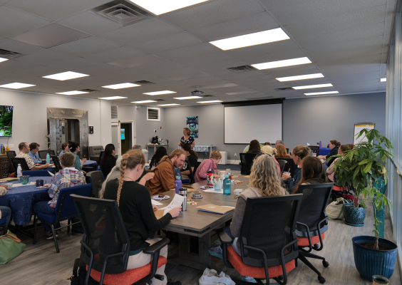 Adults seated at desks in a classroom, facing a female speaker.