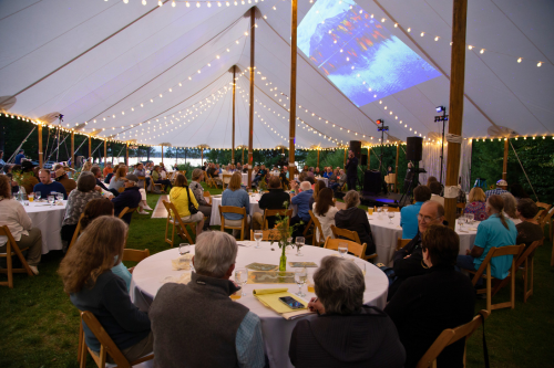 Dozens of people seated at round tables under a lit party tent.