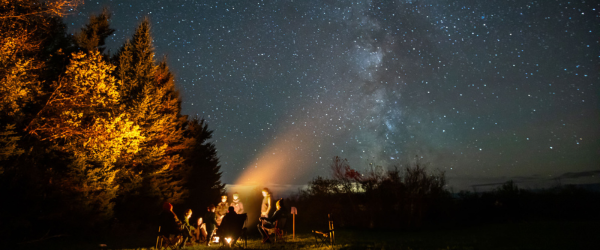 A group of people are gathered around a bonfire with the Milky Way lit up across the sky.