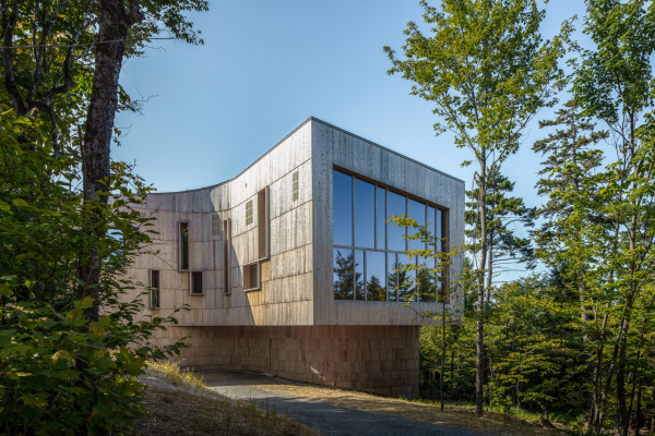 A curved exterior wall of a modern, cedar-clad building is set against a bright blue sky with trees in the foreground.