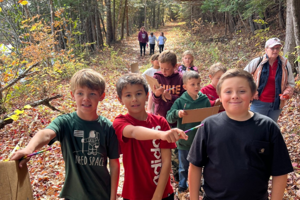A group of children walk toward the camera, smiling, on a wooded path with fall foliage.