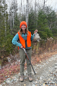 A young white woman holds a hunting rifle and a grouse on a wooded gravel road.