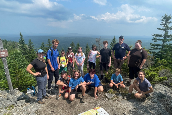 A group of teens in hiking clothes at the summit of a rocky mountaintop.