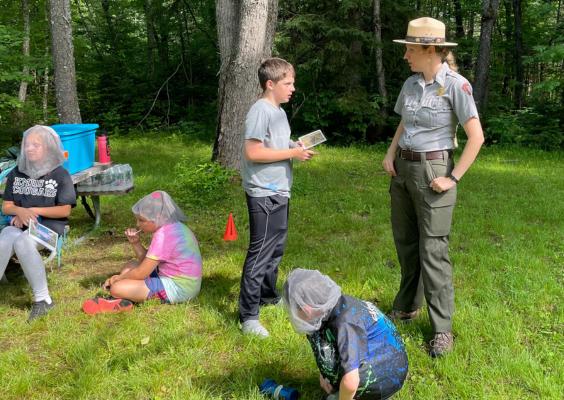 A park ranger and a student converse with other students nearby, in a clearing of trees.