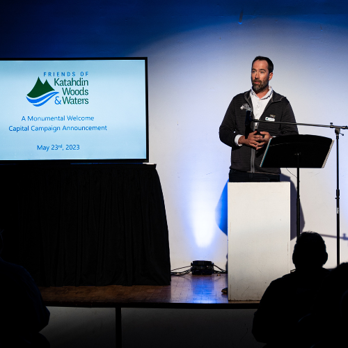 A white male stands in front of a podium and presentation screen.