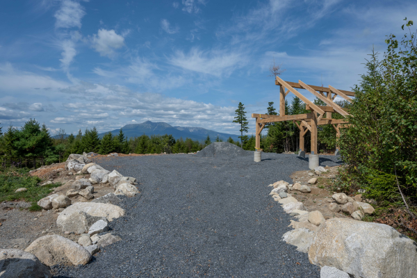 A rustic post and beam structure sits on a gravel lot with forest and mountain views s beyond.