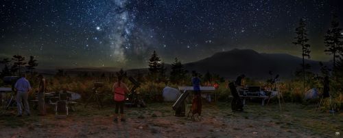 A night sky lit by the Milky Way with a mountain in the distance and telescopes and stargazers in the foreground.