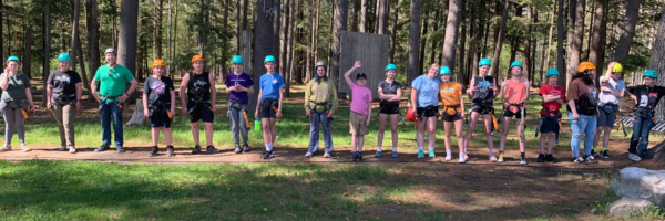 A row of white teenagers in blue helmets and climbing harnesses smile at the camera.