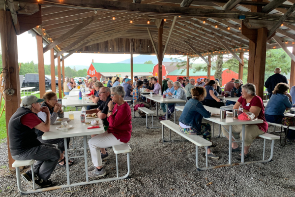 Several dozen people seated in an open air pavillion at picnic tables.