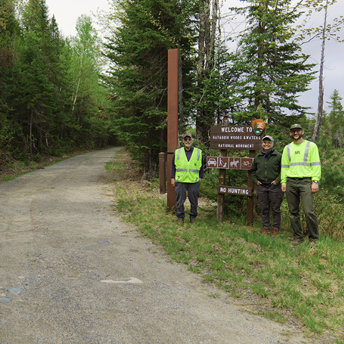 Three adults stand next to a NPS welcome sign and an open gate on a gravel road.